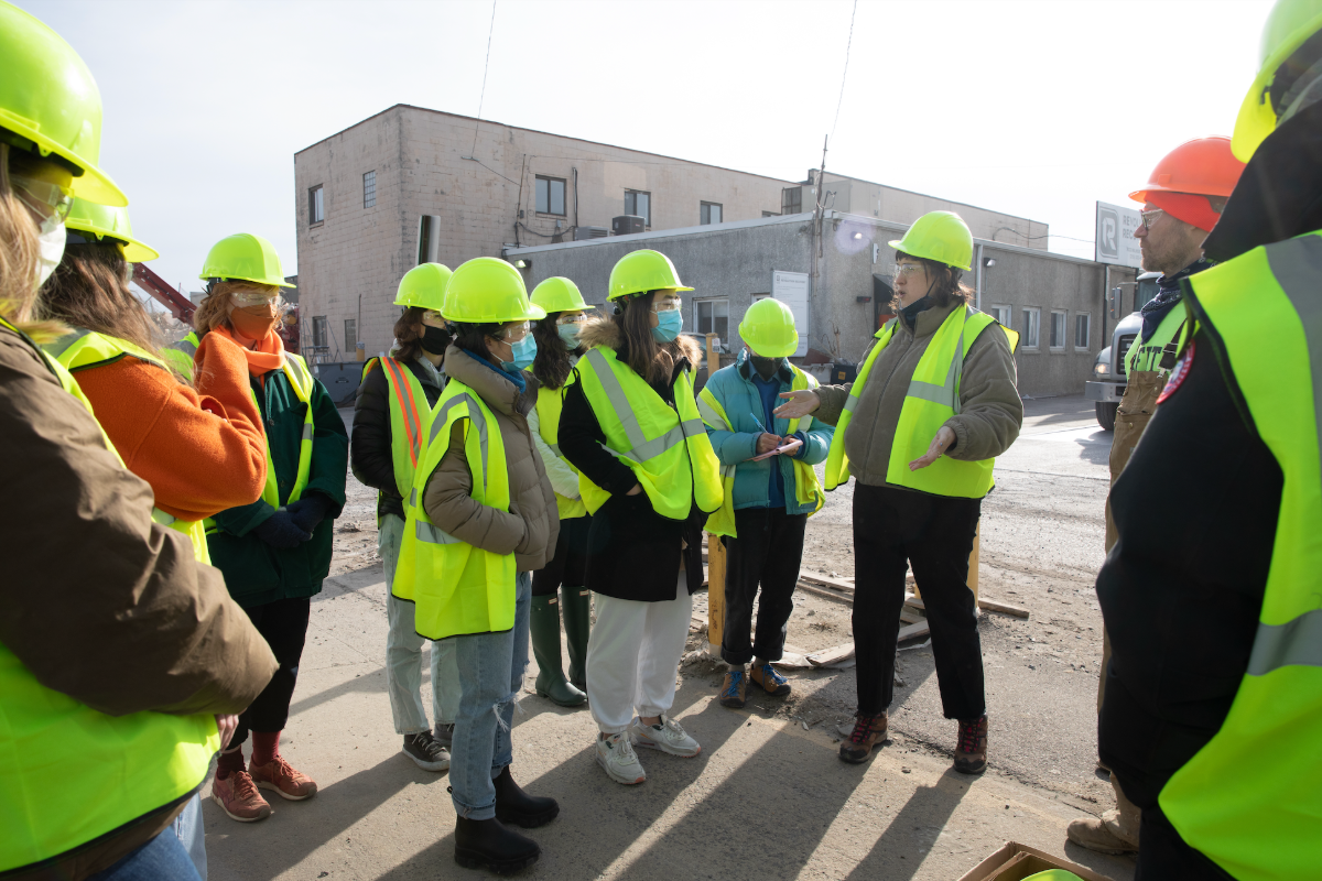 A group of people wearing yellow hard hats and vests stand near a demolition site.